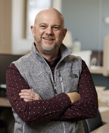 Physician Michael Sullivan, smiling with shaved head and wearing a heather grey vest over long sleeve burgundy shirt